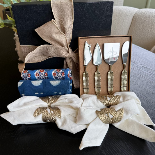 Set of four silver cheese knives in a decorative box with a white napkin and blue dishtowels on a dark surface.