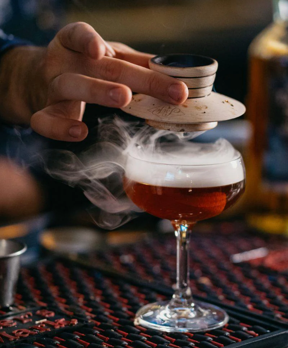 A person's hand lifting a maple wood smoking saucer from a glass of cocktail, causing smoke to emit, set on a bar mat.